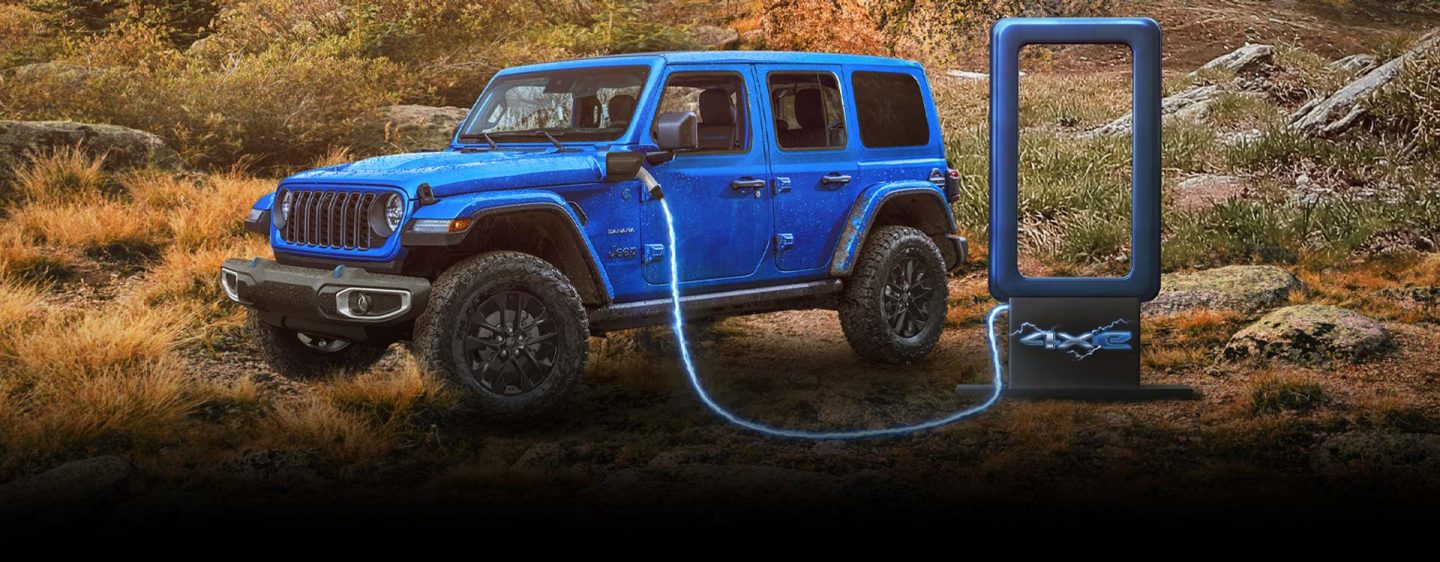 Un Jeep Wrangler Rubicon 4xe 2025 plateado, con las puertas desmontadas, desciende una colina mientras atraviesa un arroyo. Se dibujan montañas en el fondo. 4xe.