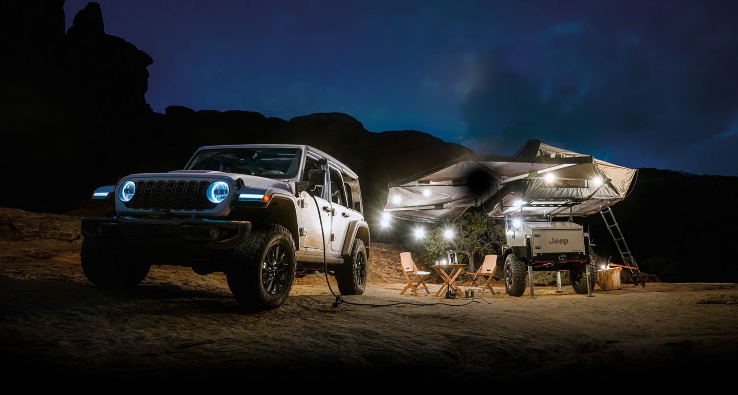 Un Jeep 4xe 2025 estacionado junto a un campamento por la noche, con un extremo de un cable inversor conectado al puerto de carga del vehículo y el otro enchufado al equipo de campamento. El camping dispone de una autocaravana marca Jeep equipada con toldos y luces.
